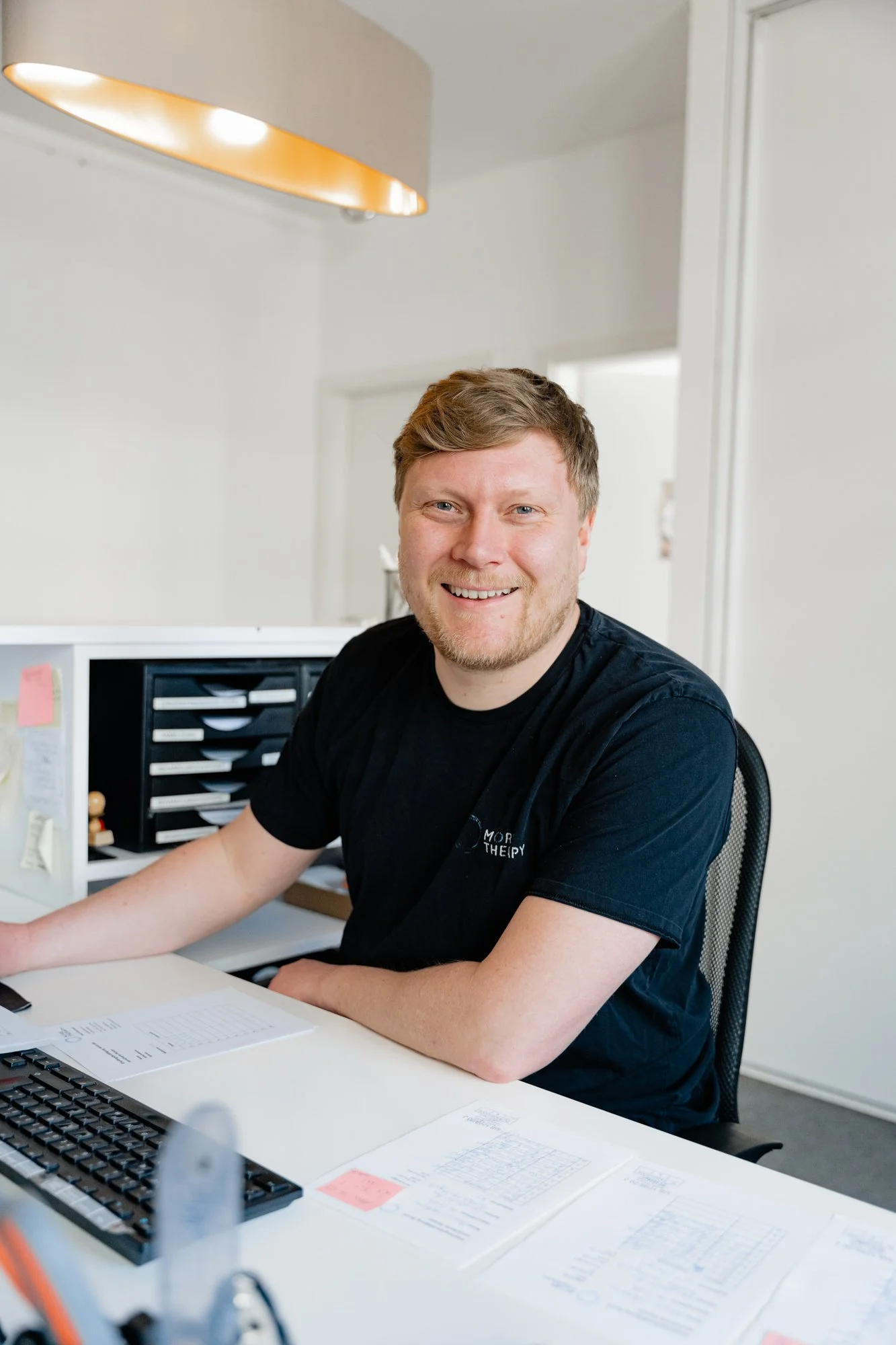 Ein Mann sitzt an einem Büroarbeitsplatz, lächelt und trägt ein schwarzes T-Shirt mit Aufdruck. Auf dem Schreibtisch sind Papiere, eine Tastatur und ein Monitor.