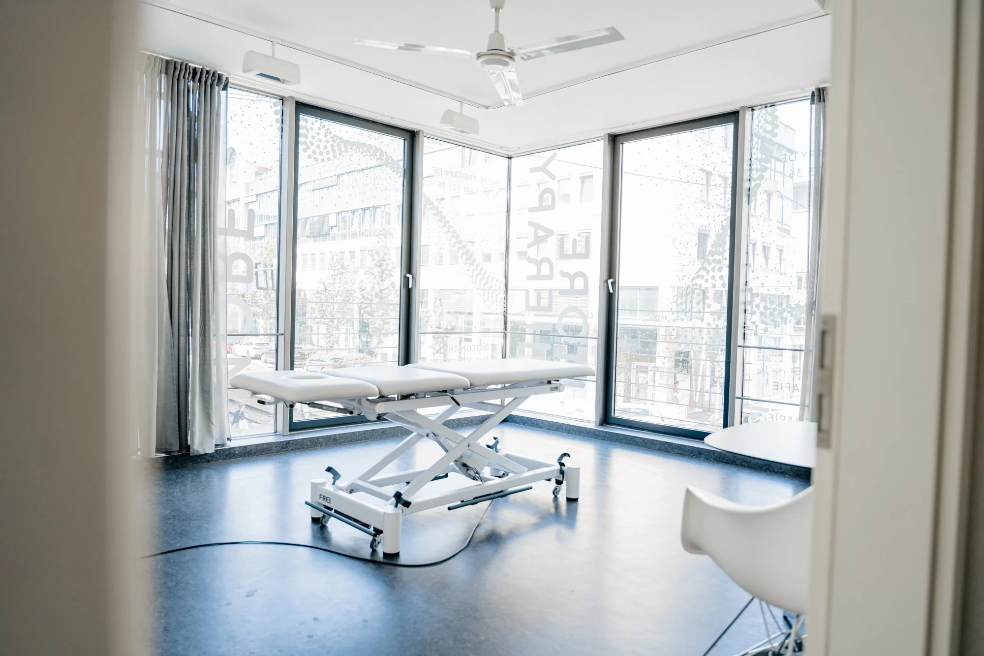Ein modern ausgestatteter Behandlungsraum, mit einem weißen Behandlungstisch in der Mitte und großen Fenstern im Hintergrund, die viel Tageslicht hereinlassen. Osteopathische Behandlung Stuttgart