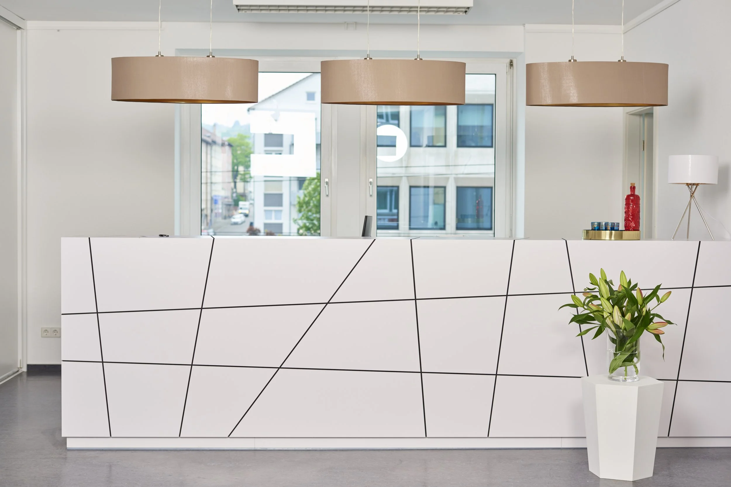 Modernes Empfangs- oder Rezeptionstresen in einem Büro mit weißen Flächen und schwarzen Linien, dekoriert mit einer Vase mit weißen Lilien, im Hintergrund Fenster, Brown-Lampenschirme und eine Stehleuchte. MORE Therapy