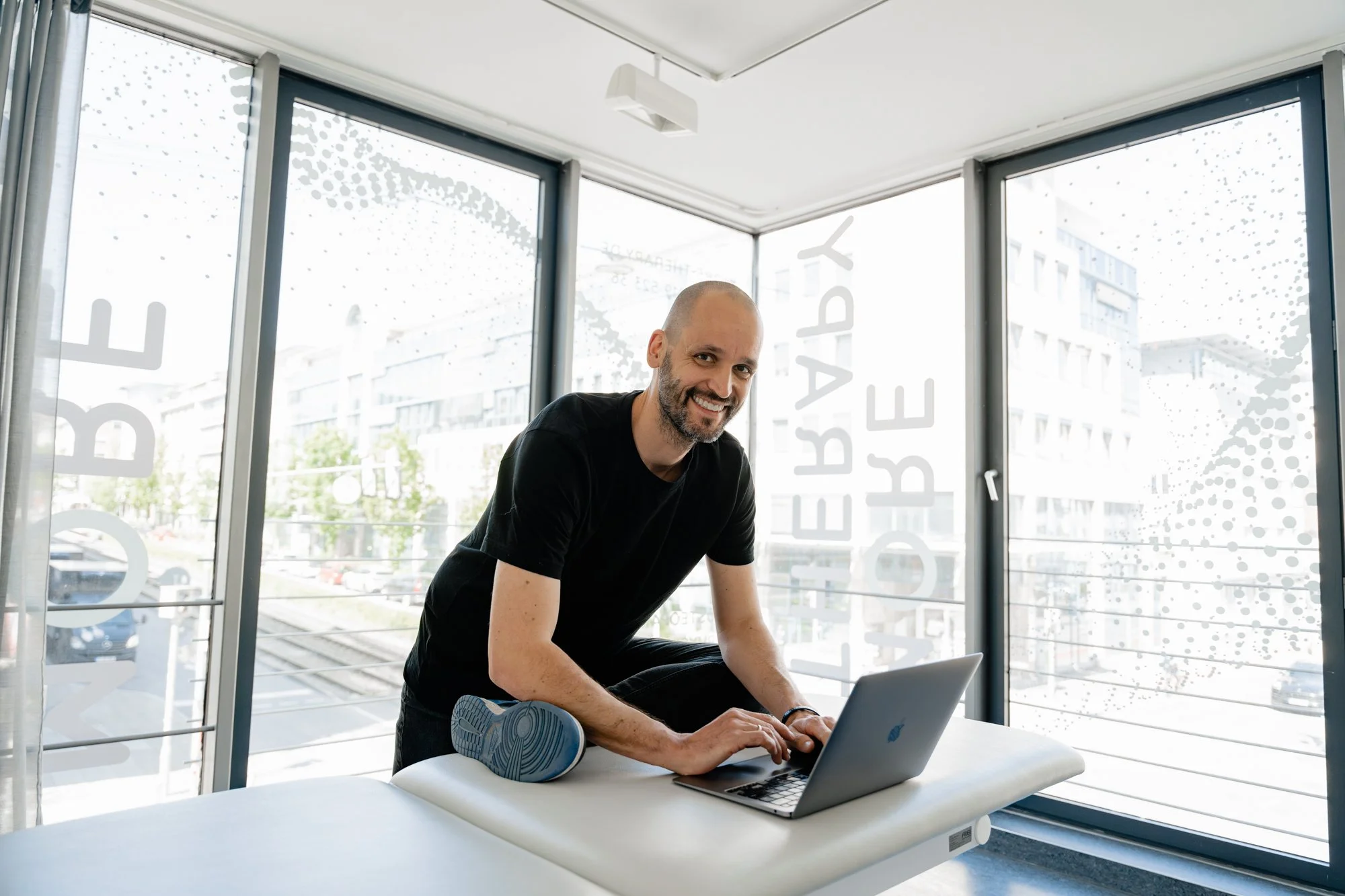 Mann trainiert im Fitnessstudio, legt seine Fußsohle auf eine Gymnastikmatte, während er an einem Laptop arbeitet, im Hintergrund Fenster mit Sicht auf ein Gebäude