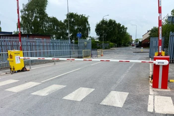 Closed railway crossing with barrier gate, warning lights, and gate control box on the left, pedestrian crossing lines on the road, surrounded by fence and trees in the background.