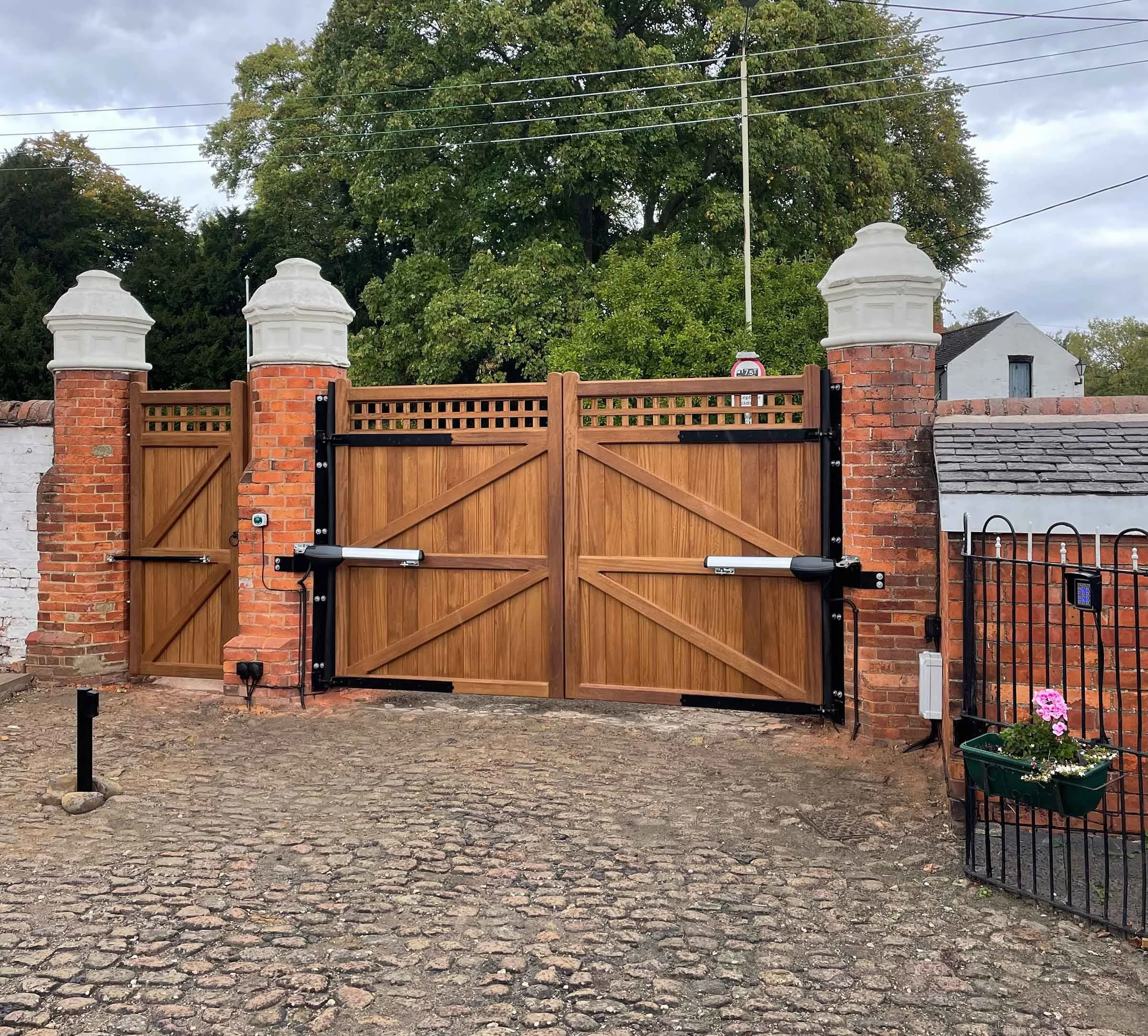 A wooden gate with brick pillars on either side, topped with white decorative caps, set against a backdrop of green trees and a cloudy sky.