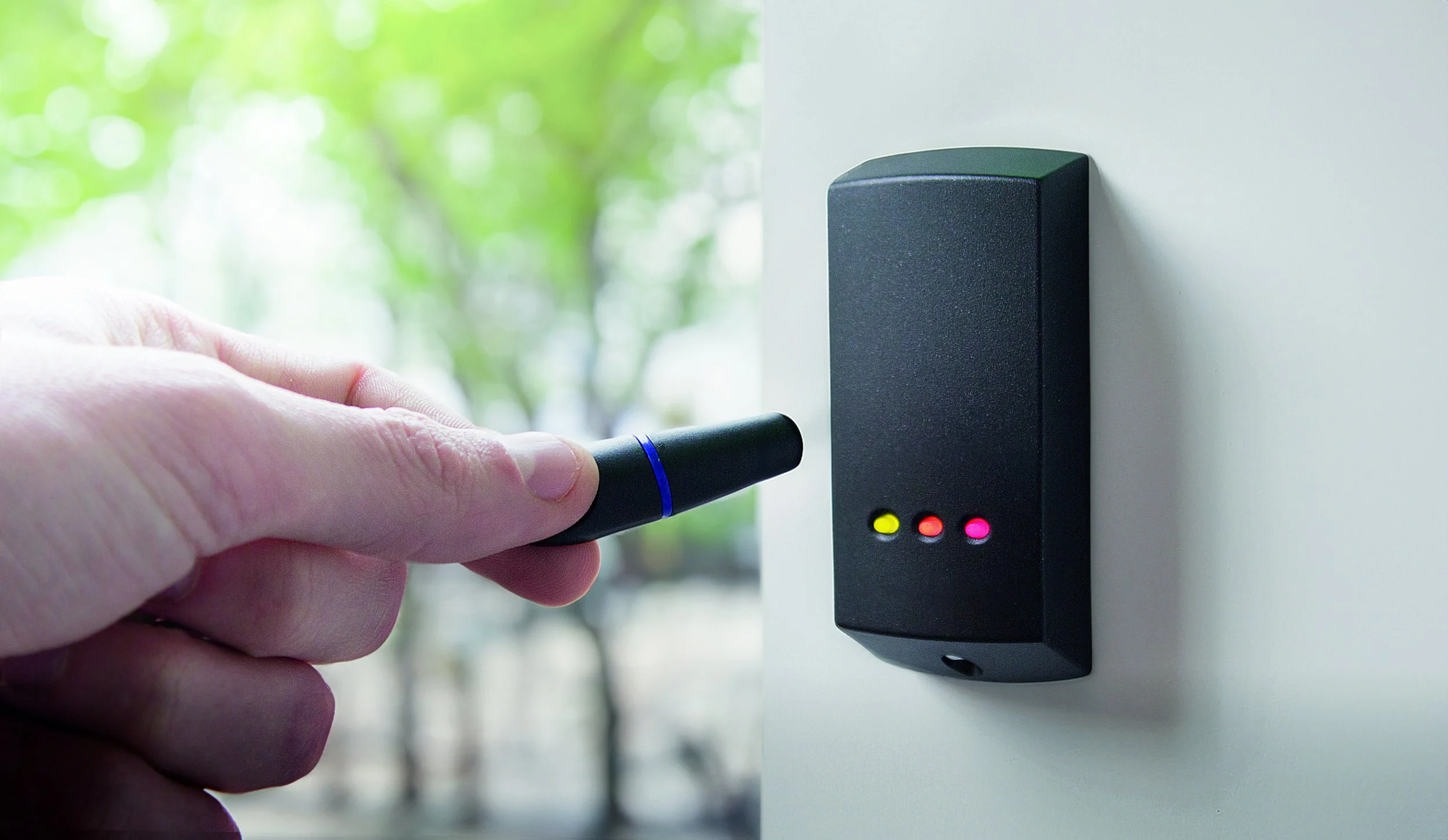 Close-up of a hand holding a black RFID key fob near an electronic access control panel with three indicator lights, one yellow, one red, and one pink, mounted on a white wall.