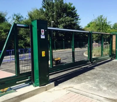 A long green metal fence with a sliding gate, situated on a paved area near trees and greenery in the background.