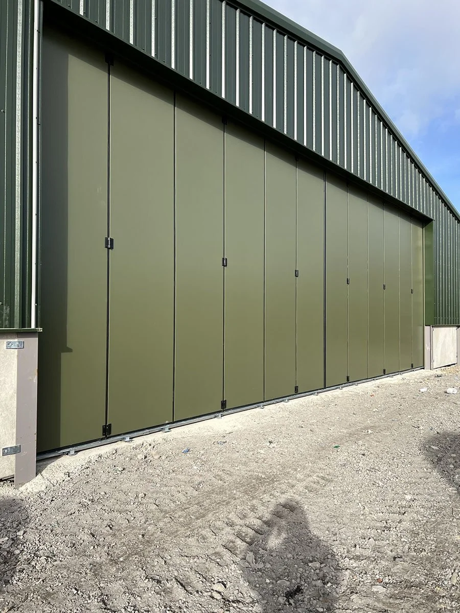 Green industrial building with large sliding doors on a gravel surface, under a partly cloudy sky.