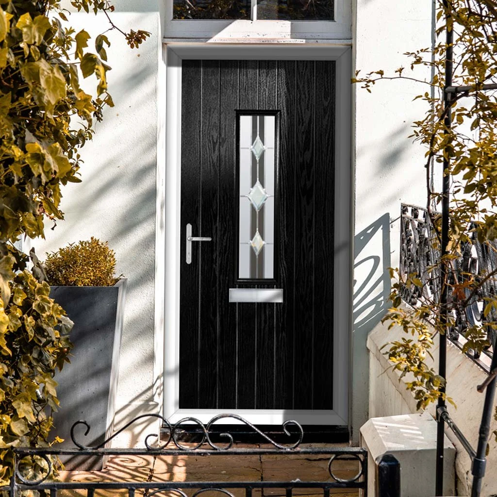 A black front door with white frame, a decorative glass panel in the center, a mail slot, and a silver handle, framed by white walls with plants on both sides.