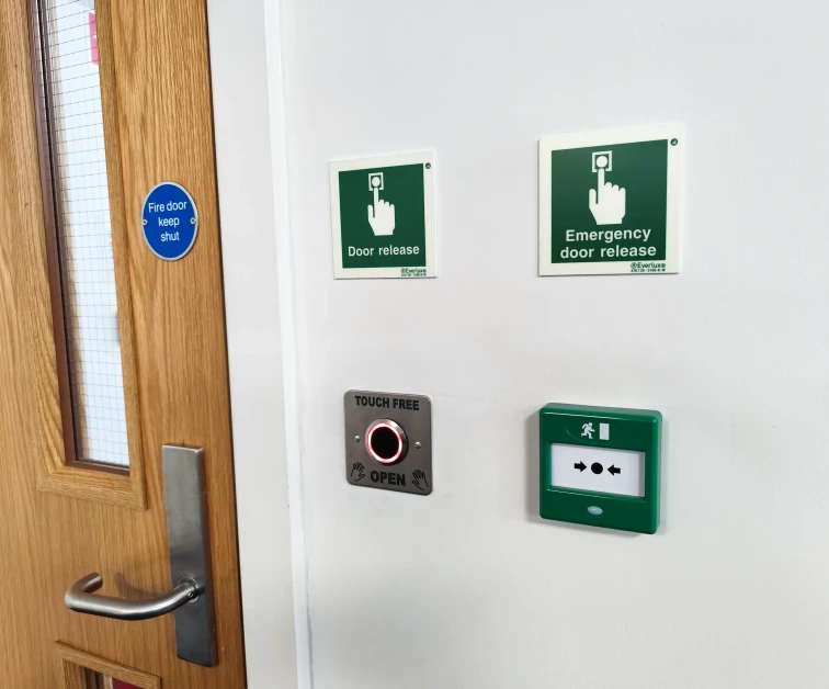 Wall with emergency and door release signs, a touch-free door opener button, and a fire door keep shut sign on a wooden door.