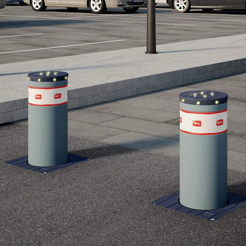 Two cylindrical parking lot barriers with rounded tops, lights along the edges, and BFC logos on white bands, situated on a paved sidewalk near a parking lot.