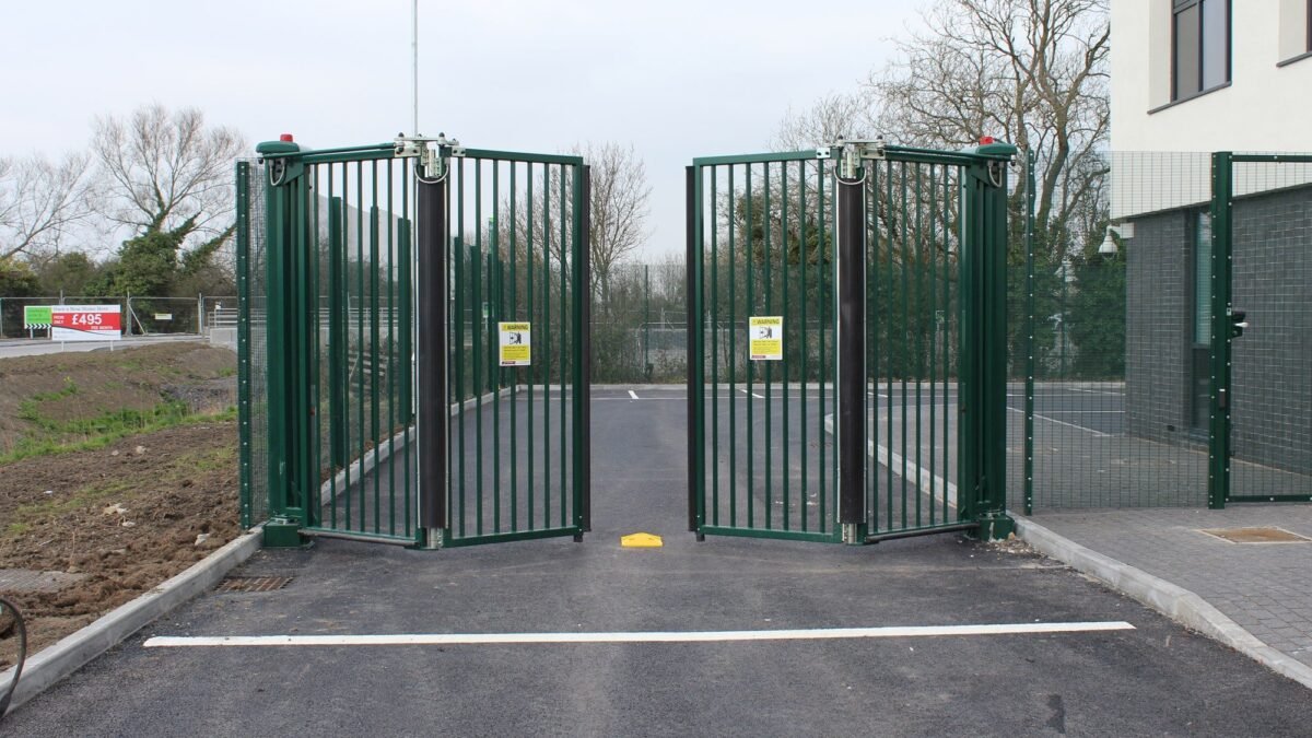 A green metal gate with automatic sliding mechanisms at a parking lot entrance, flanked by a building on the right and open land on the left, with trees in the background.