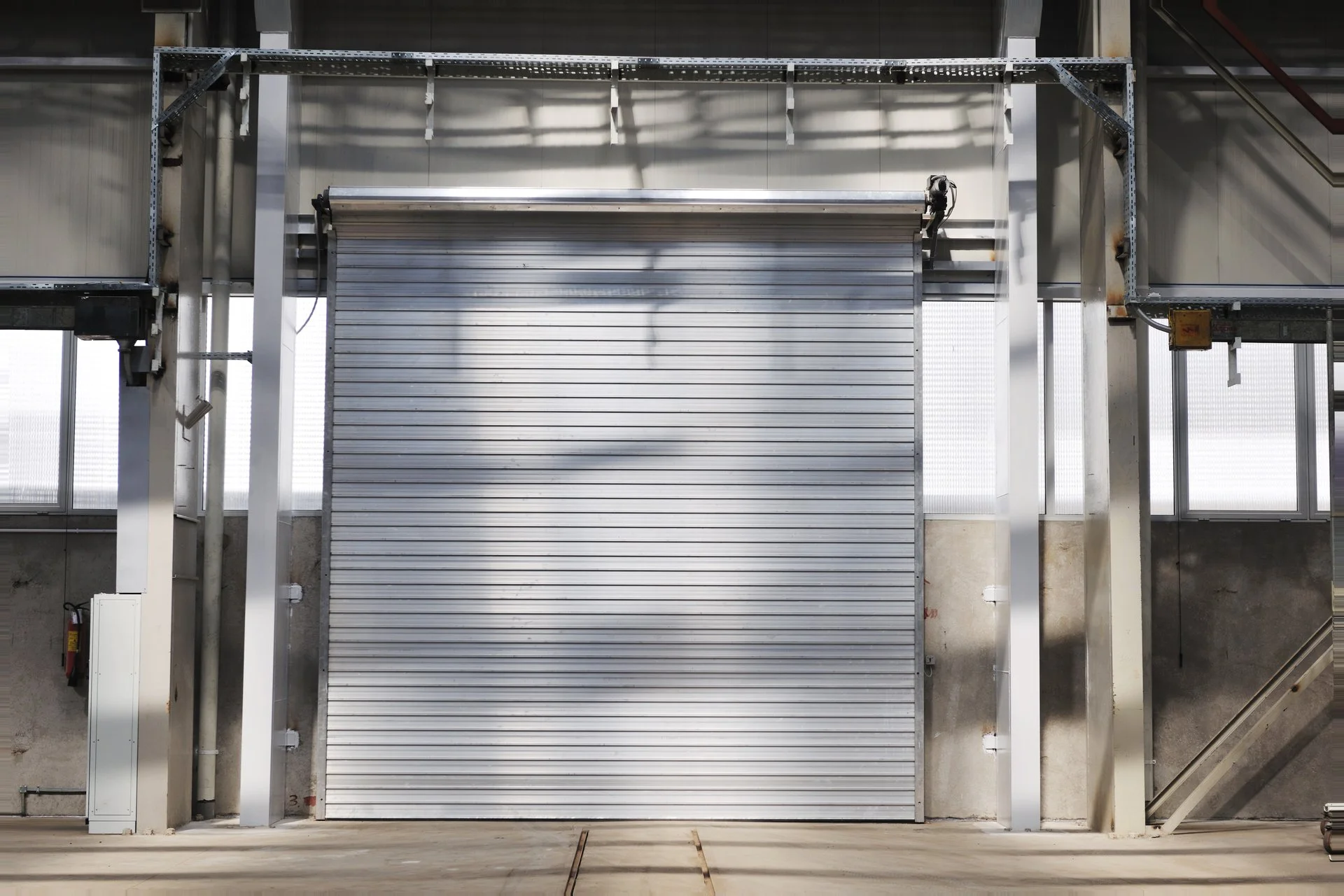 Industrial indoor space with a metallic roll-up door, supported by structural steel beams and piping, with a concrete floor and some machinery and equipment partially visible.