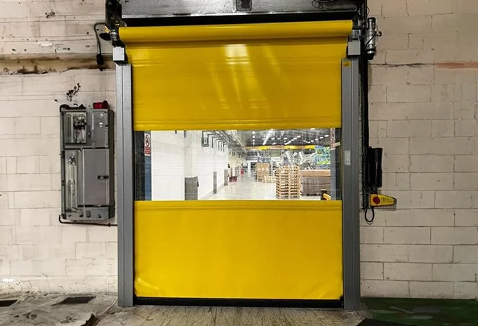 Yellow industrial roll-up door leading to a warehouse interior with pallets and stacked boxes.
