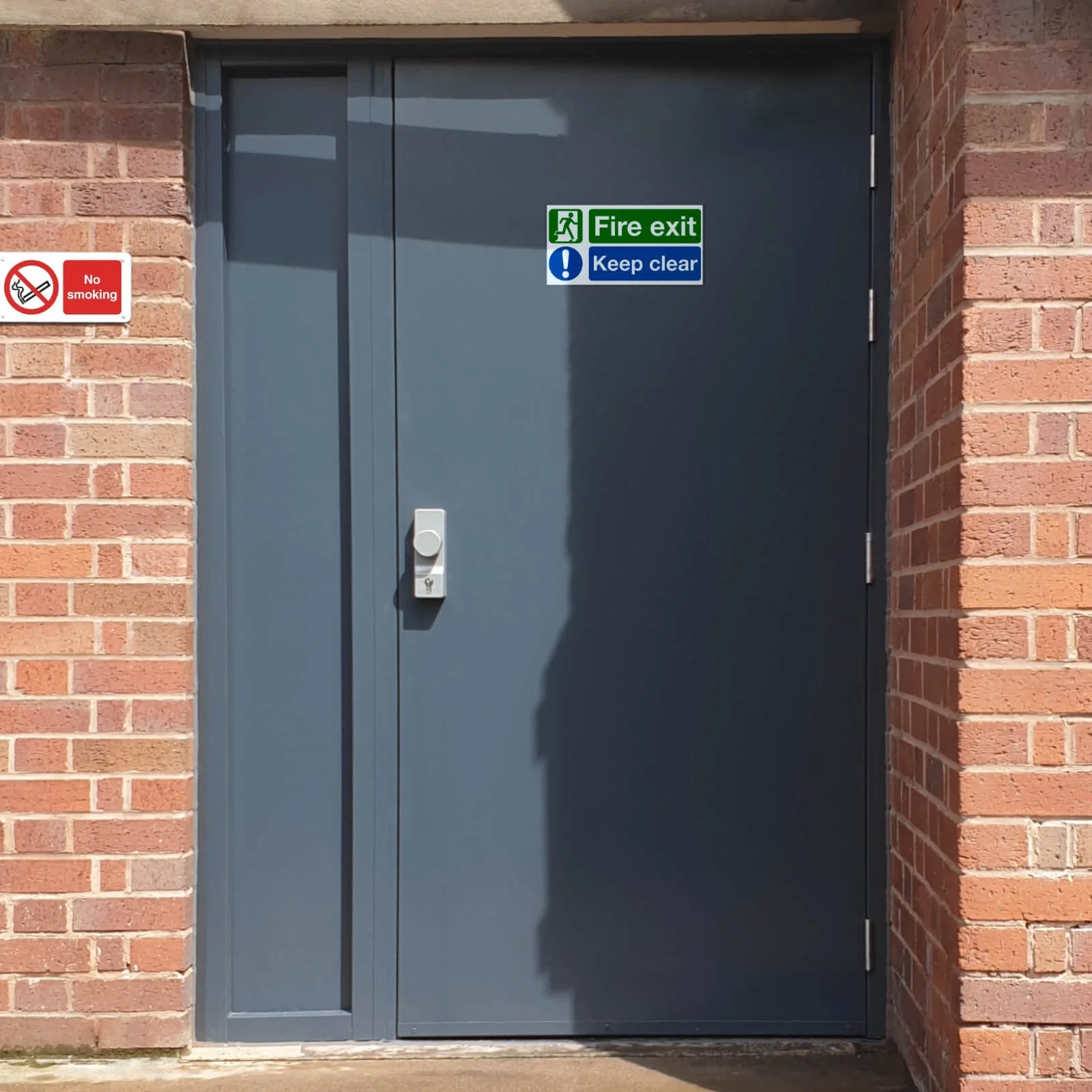 Metal door with smoke and fire exit signs, set in a brick wall with no smoking sign nearby.