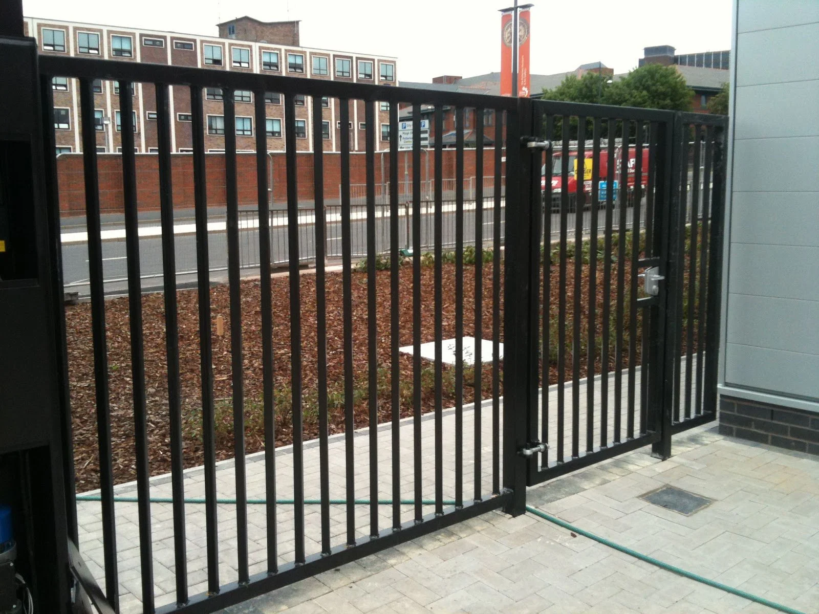 Black metal gate and fence outside of a building, with a view of a street, trees, and buildings in the background.