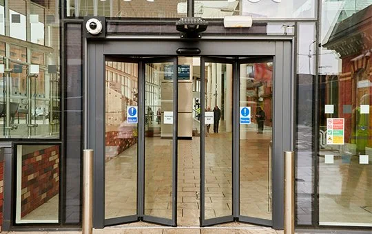 Automatic sliding glass entrance door with security camera above, located in a building lobby.