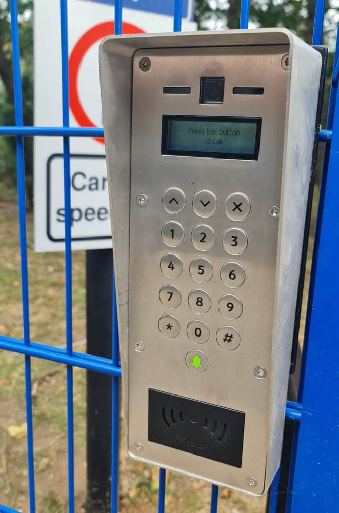 A metal security keypad with a small display screen, number pad, and RFID reader mounted on a blue fence. The display shows "Press bell button to call."
