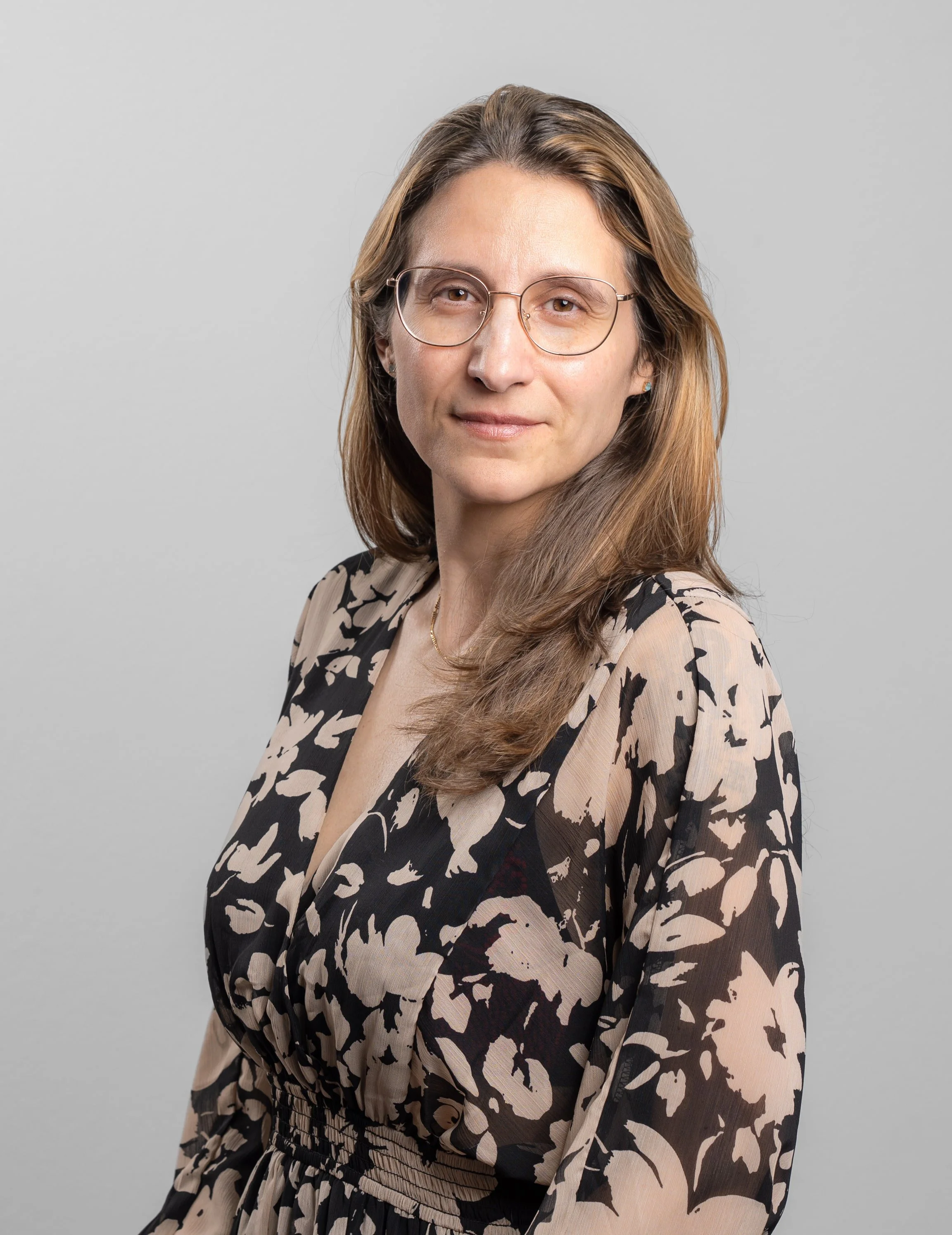 Porträt einer Frau mit Brille, braunen Haaren und einem schwarz-beigen Musterkleid, vor hellem grauen Hintergrund.