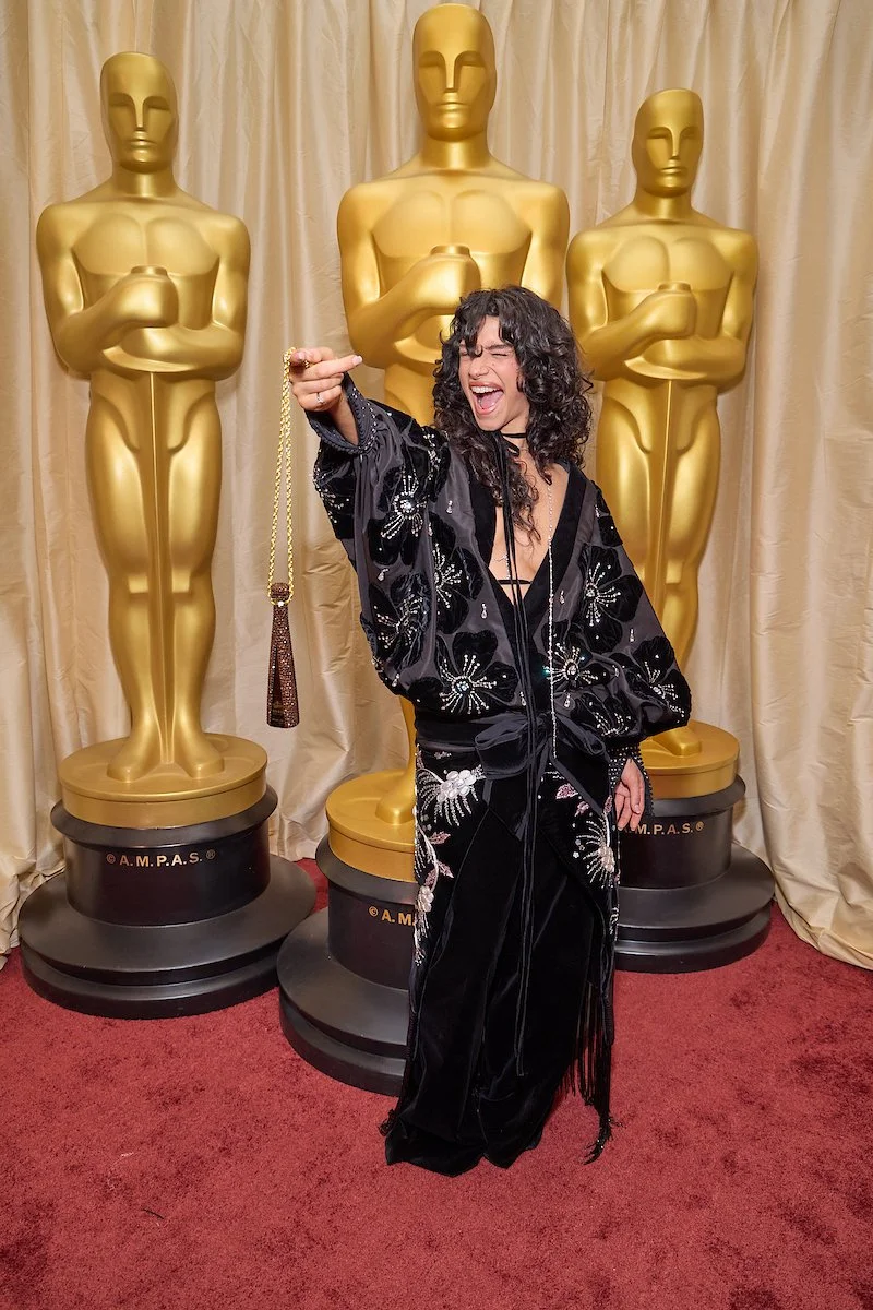 A woman with curly black hair in a black, embellished outfit makes a rock and roll gesture in front of three large, gold Oscar statues at an awards ceremony.