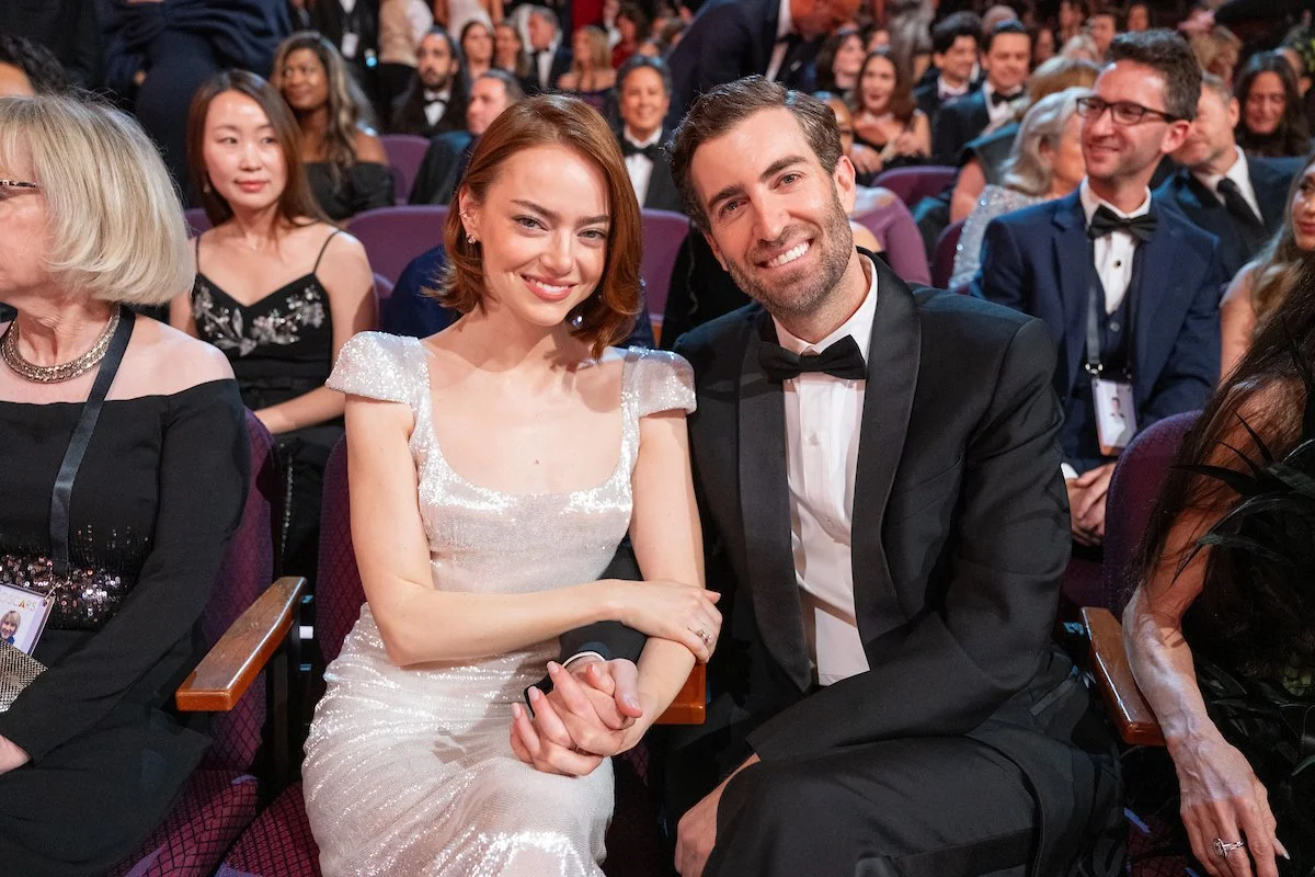 A man and woman in formal attire sitting in an audience at an event, holding hands and smiling at the camera. The woman is wearing a sparkly white dress and the man a black tuxedo with a bow tie.
