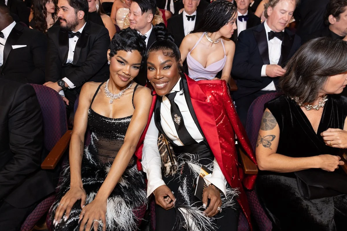 Two women sitting next to each other at a formal event, smiling at the camera, surrounded by well-dressed men and women in tuxedos and evening gowns.