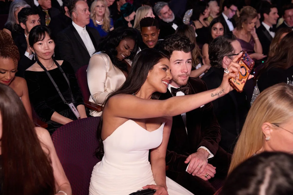 People attending an event, with a woman in a white strapless dress taking a selfie with a man in a tuxedo, surrounded by seated attendees in formal attire.
