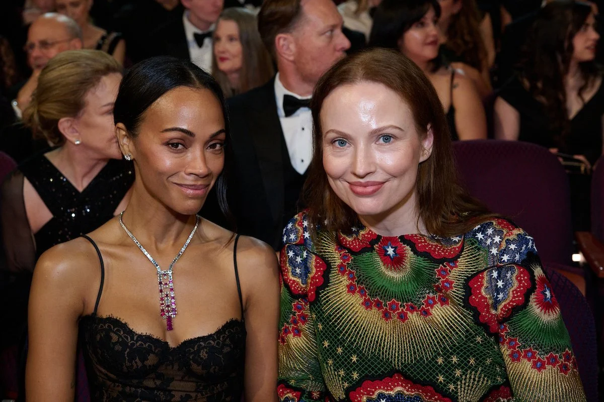Two women sitting in an audience at a formal event, smiling at the camera. The woman on the left has dark hair, medium skin, and is wearing a black lace dress and a diamond necklace. The woman on the right has light skin, red hair, and is wearing a c
