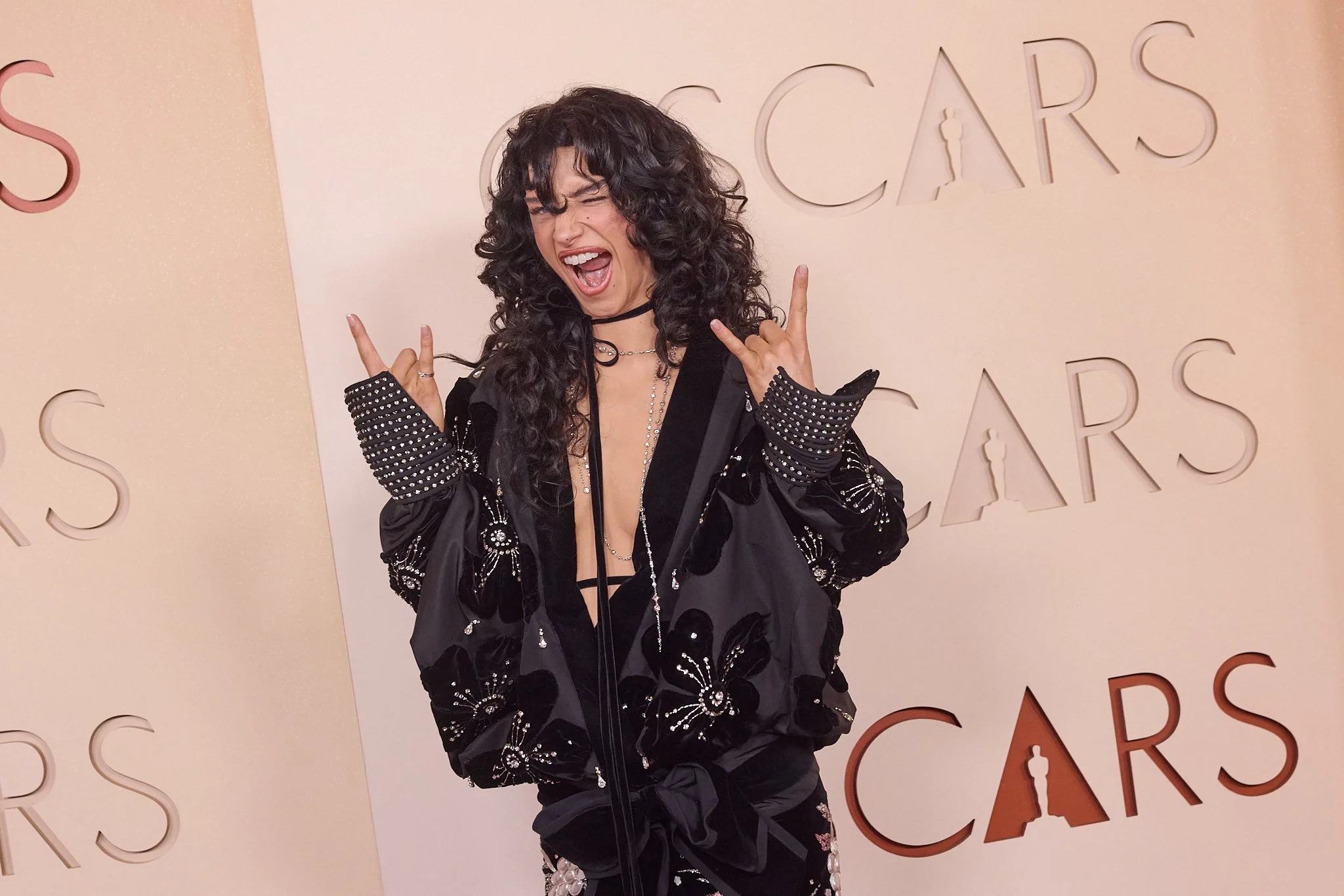A woman with curly dark hair, wearing a black floral jacket and jewelry, making a rock hand gesture and smiling with her eyes closed at a Cannes event.