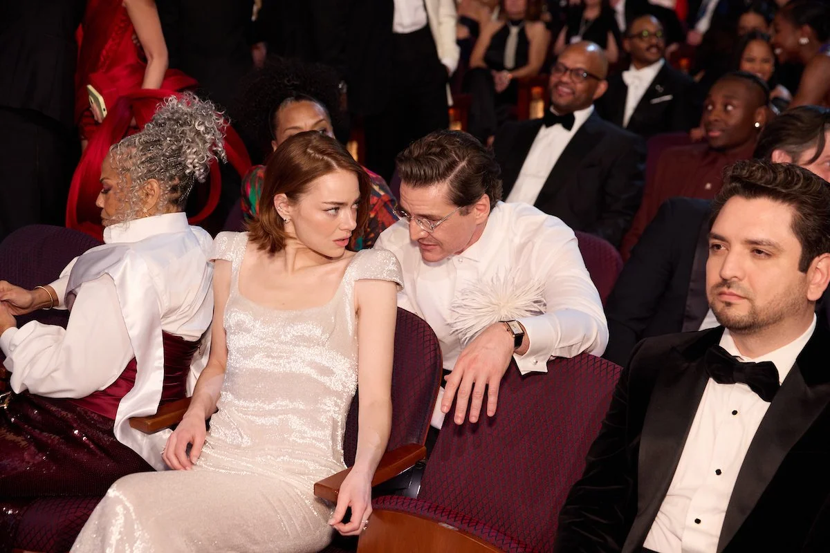 A woman in a light-colored dress and a man in a white shirt are seated close together, engaged in a conversation at a formal event. The woman has short auburn hair, and the man is wearing glasses. Other guests in formal attire are visible in the back