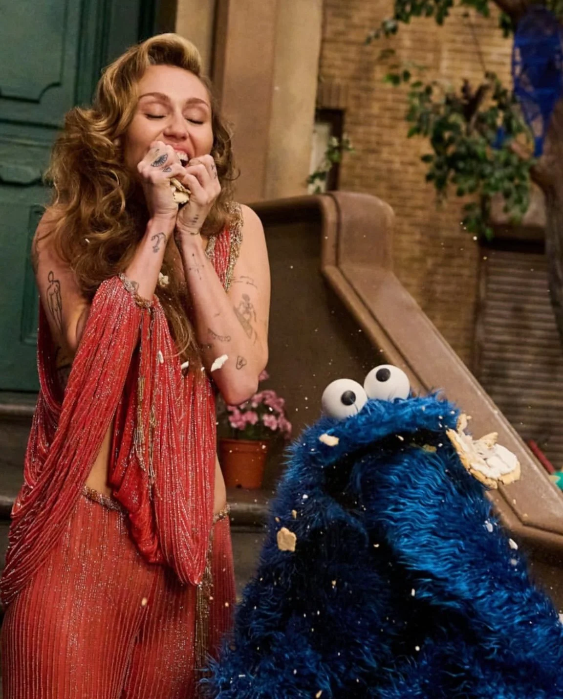 A woman with long curly red hair and tattoos on her arms is eating a cookie, smiling with her eyes closed. She is wearing a red, sleeveless, shiny dress. A large blue furry puppet resembling Cookie Monster from Sesame Street is sitting nearby, with cookie crumbs on its face and body, and googly eyes on top of its head. The background has brick walls, a green door, and a potted plant.