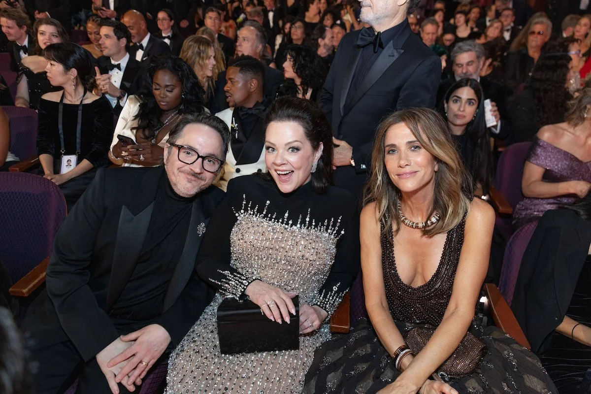 Three people seated at an event, with a large crowd in the background. The man on the left wears glasses and a black tuxedo, the woman in the center wears a black dress with pearl embellishments and holds a black clutch, and the woman on the right we