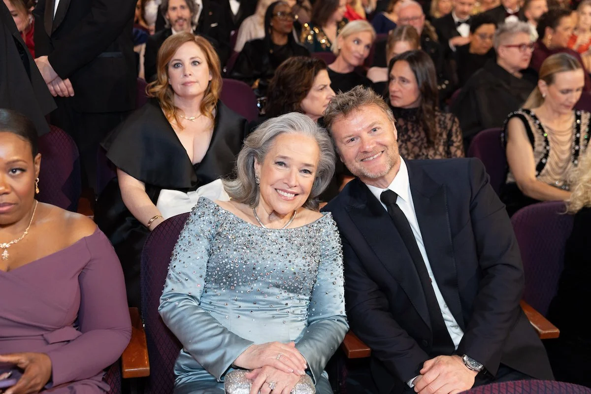 A woman and a man are sitting side by side at an event, smiling at the camera. The woman has gray hair and is wearing a sparkly, light blue dress with embellishments, while the man has light brown hair and is dressed in a black suit with a white shir