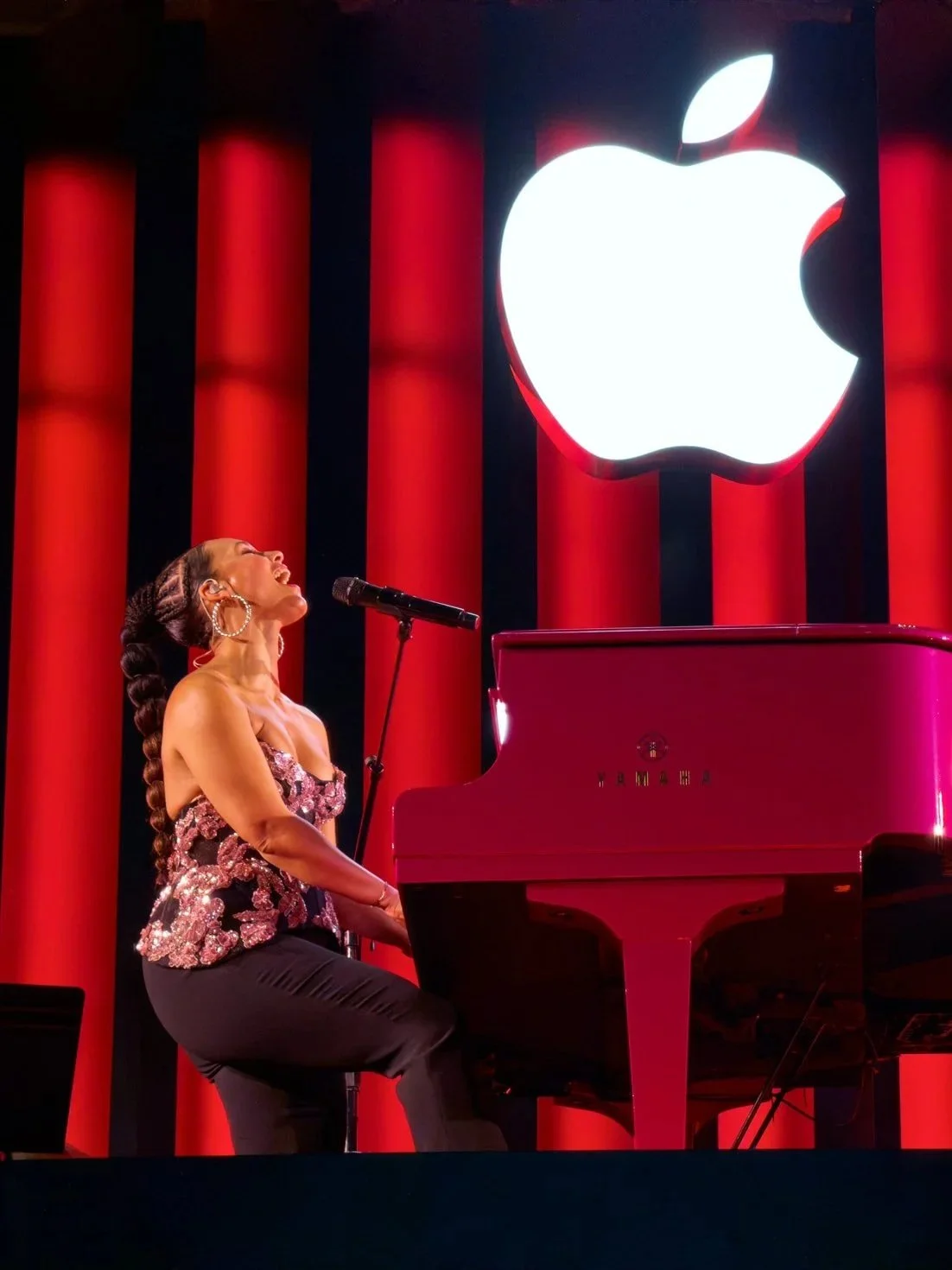 A woman singing at a piano on stage with a large illuminated Apple logo in the background.