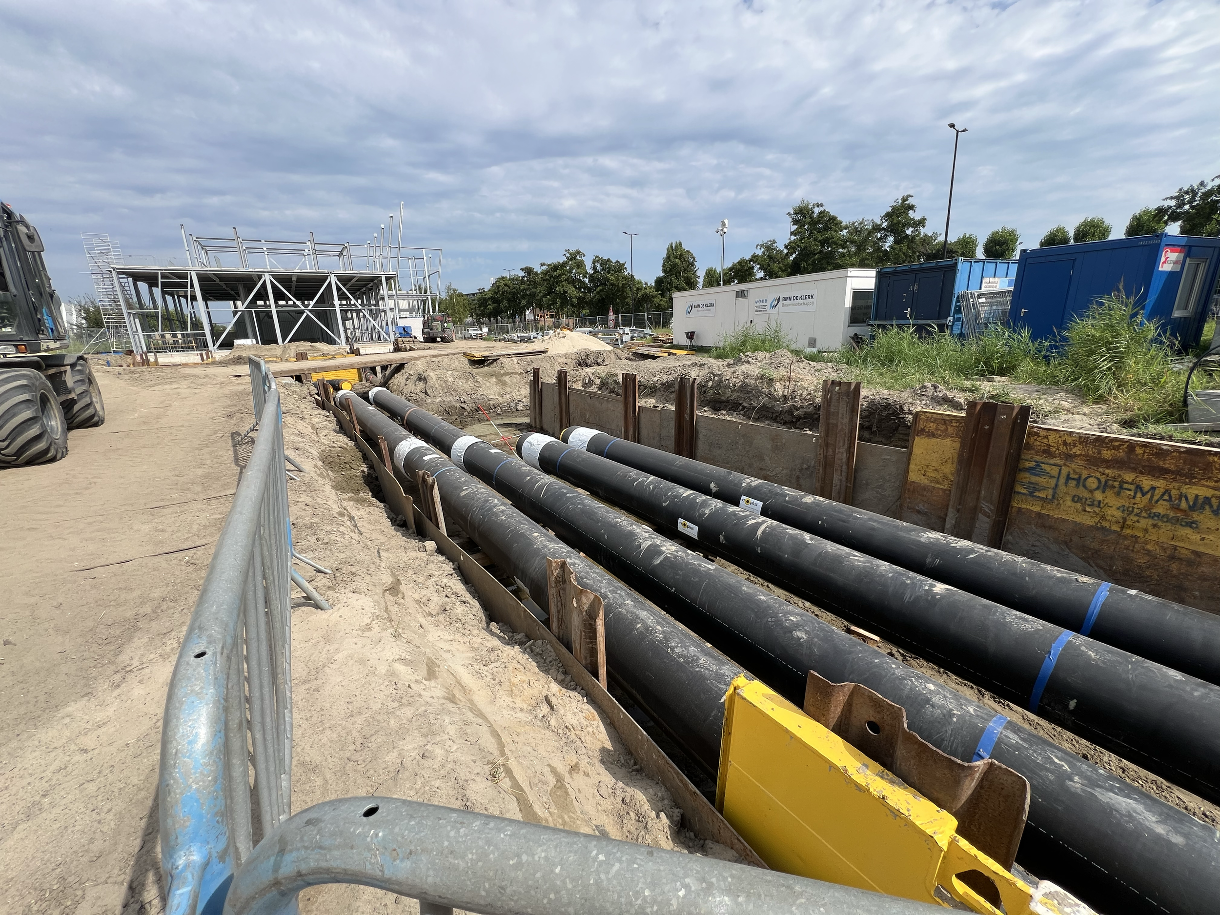 Bouwplaats met grote zwarte waterleidingen, een bouwsteiger in de achtergrond, en verschillende bouwmaterialen en voertuigen, onder een bewolkte lucht.