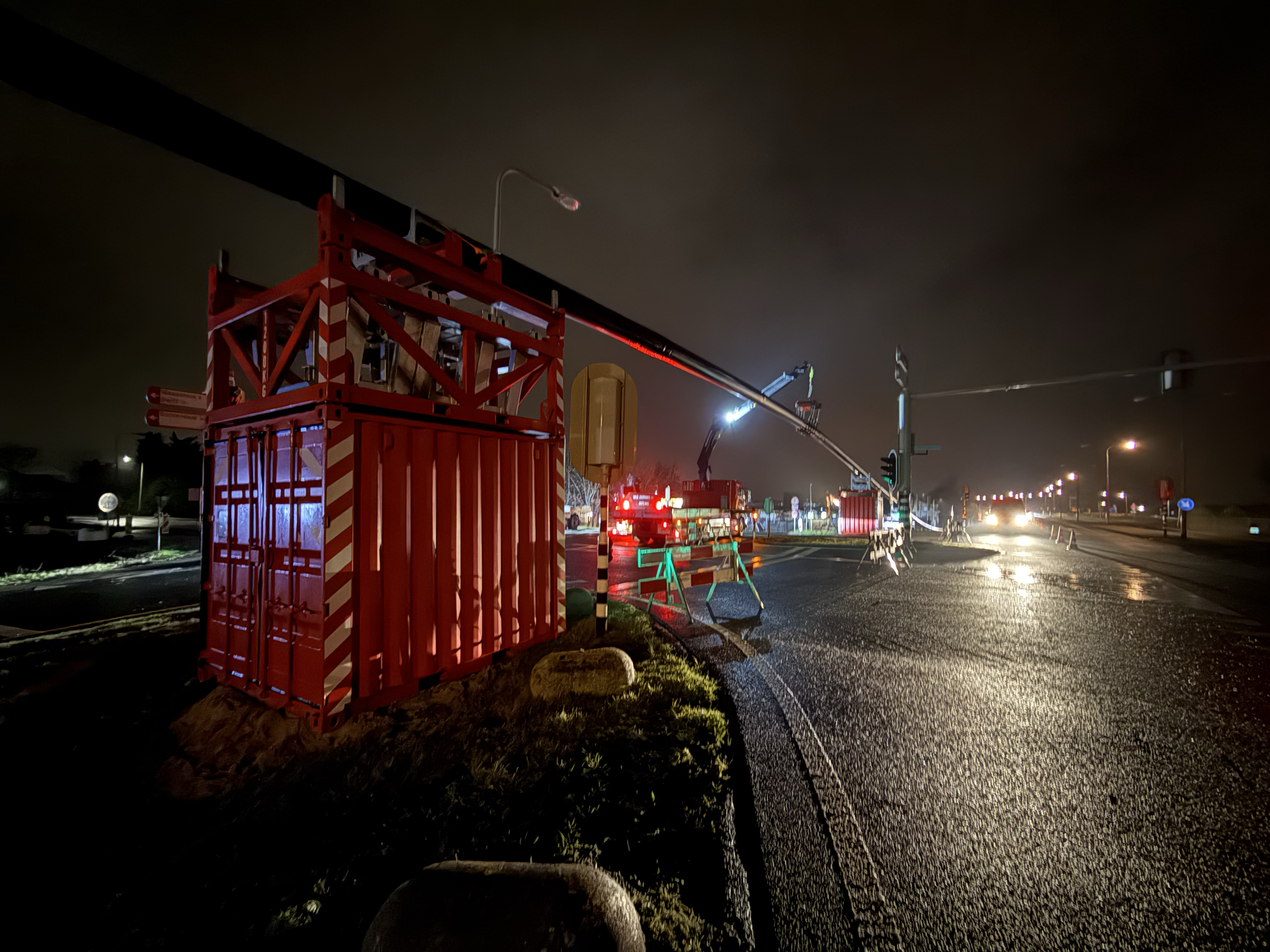 Een wegwerkersbedrijf voert nachtelijke werkzaamheden uit, met een grote kraan en steigers langs de weg. De omgeving is donker en de weg is nat, vermoedelijk na regen, met straatverlichting die het werkgebied verlicht.