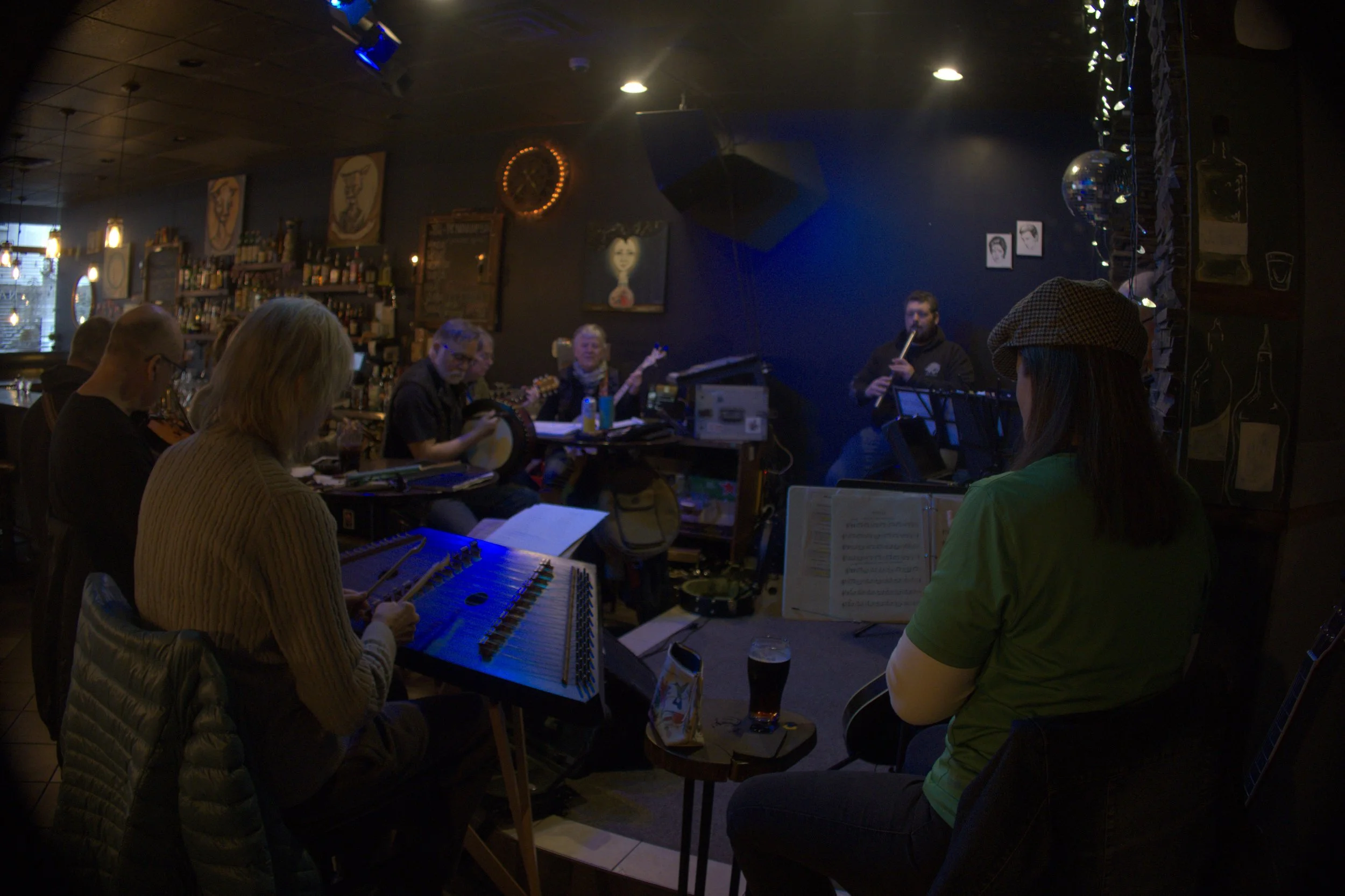 A live music performance in a dimly lit bar, featuring musicians playing instruments including a keyboard, drums, flute, and guitar, with patrons seated at tables.