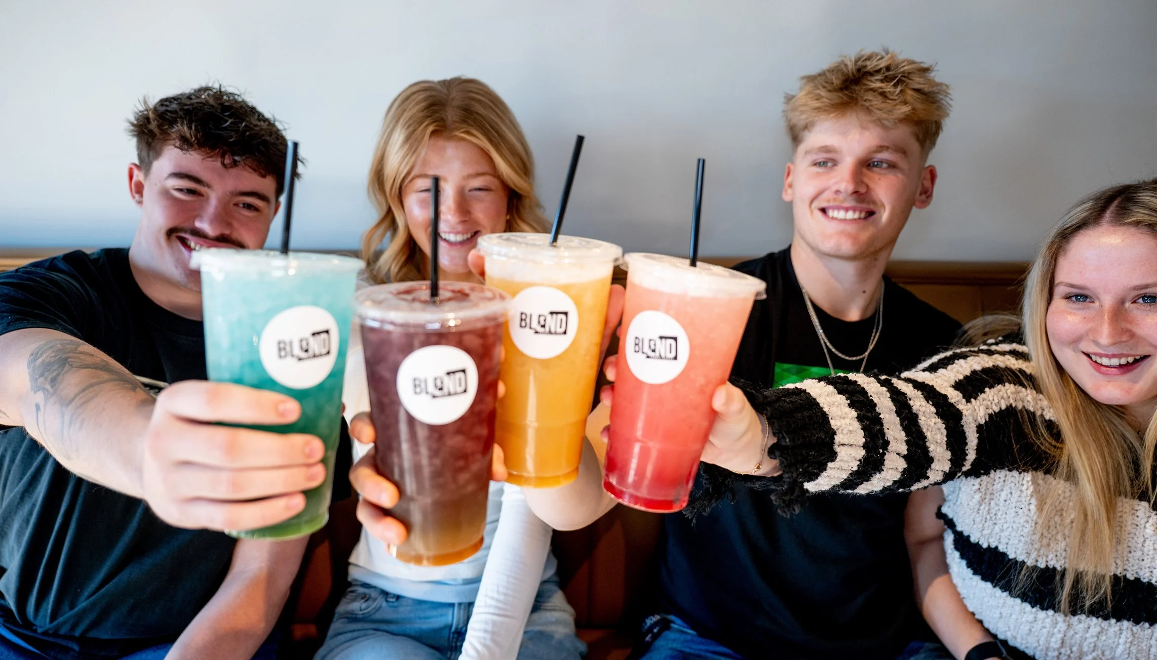 Five young adults sitting at a table holding colorful smoothies and smiling.