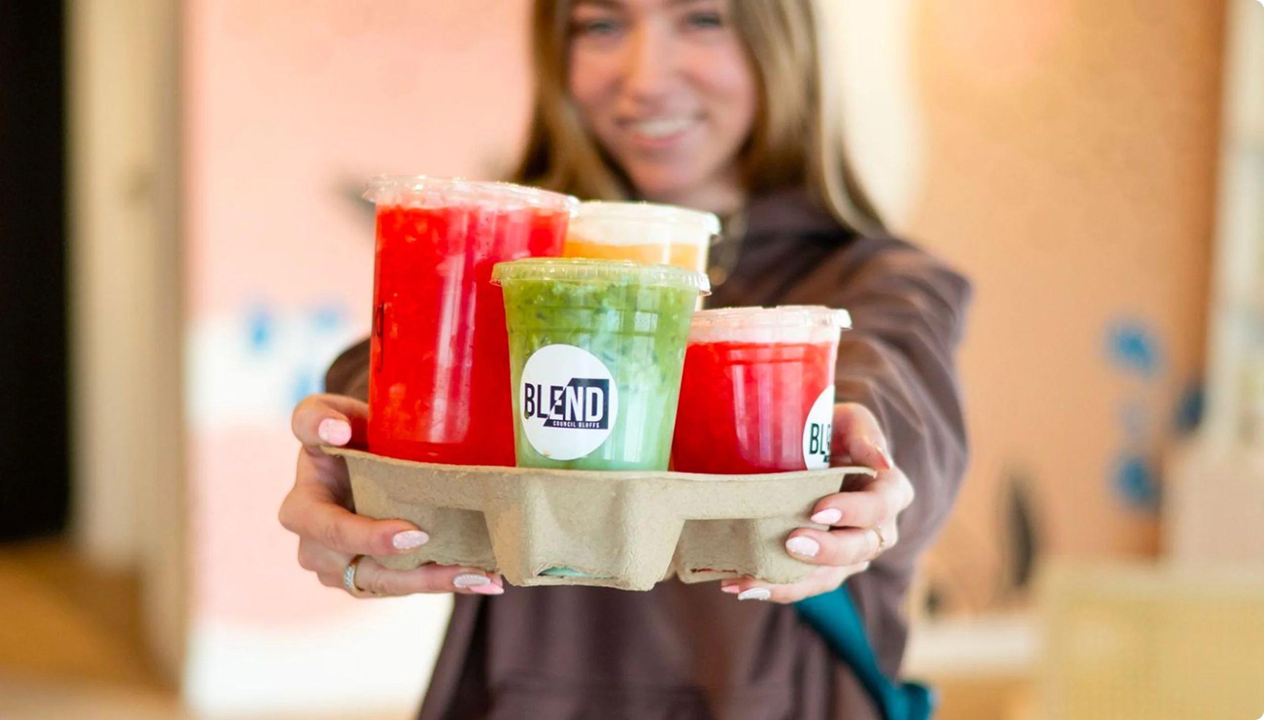 A woman holding a tray with colorful smoothies, three red, one green, and one yellow, in plastic cups with lids.