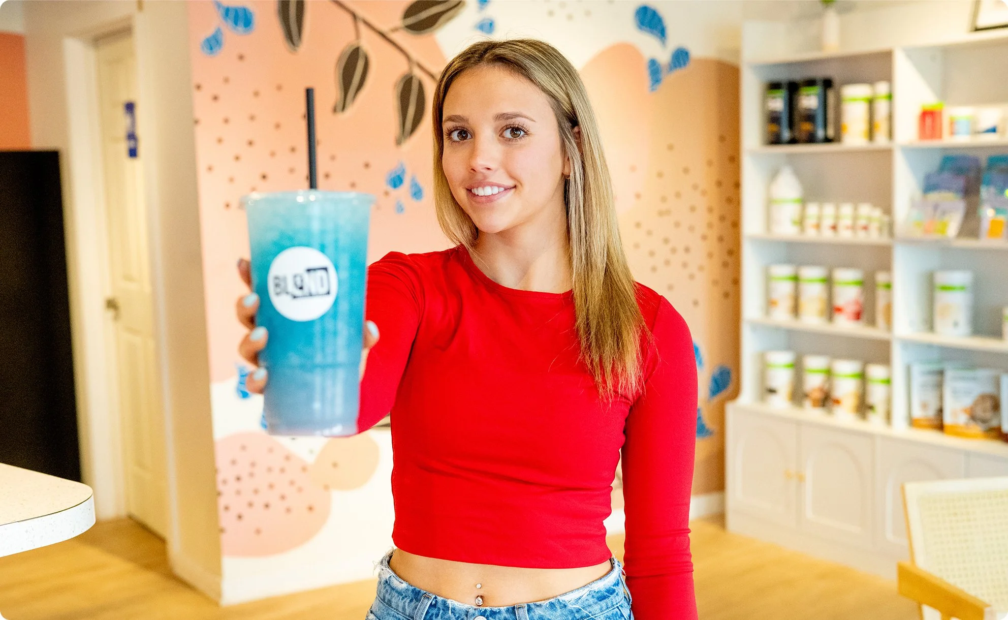 A woman in a red long sleeve top and blue jeans holds a large blue drink with a black straw, smiling at the camera inside a brightly decorated cafe or dessert shop.
