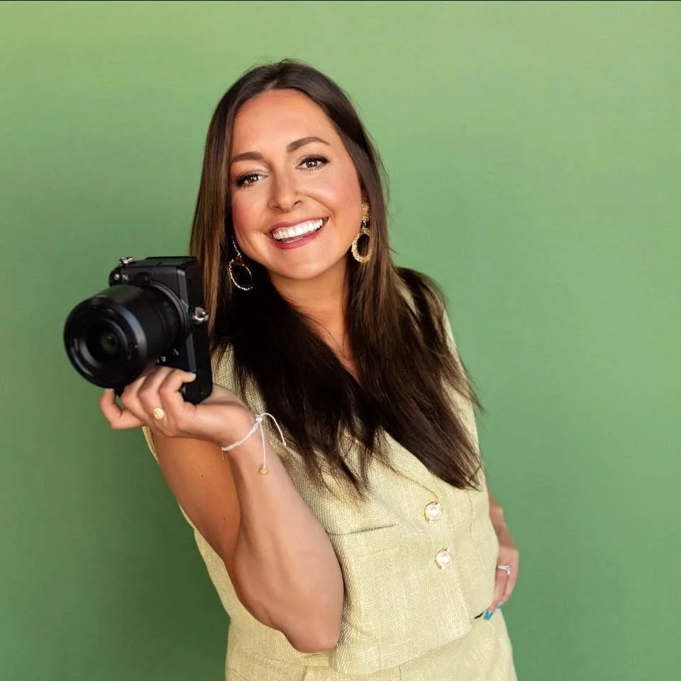 Woman with long brown hair smiling and holding a black camera, wearing gold earrings, a gold vest, and a bracelet, standing against a gradient green background.
