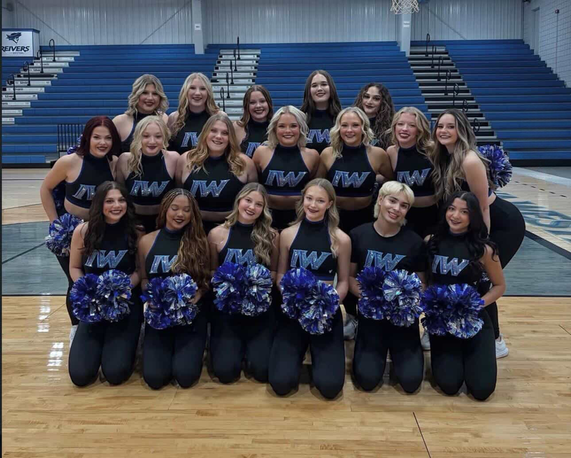 Group of young female cheerleaders in black and blue uniforms with pom-poms, posing for a team photo in a gymnasium with blue bleachers in the background.