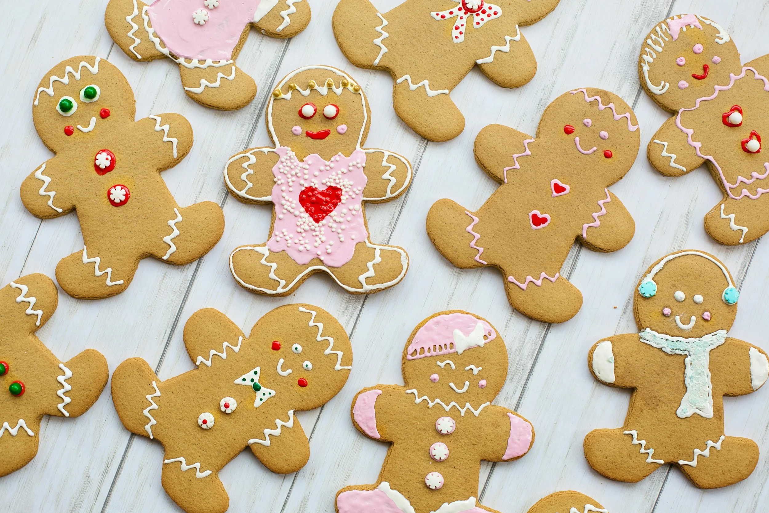 Assorted gingerbread cookies decorated with white, pink, and red icing, some with sprinkles, on a white wooden surface.