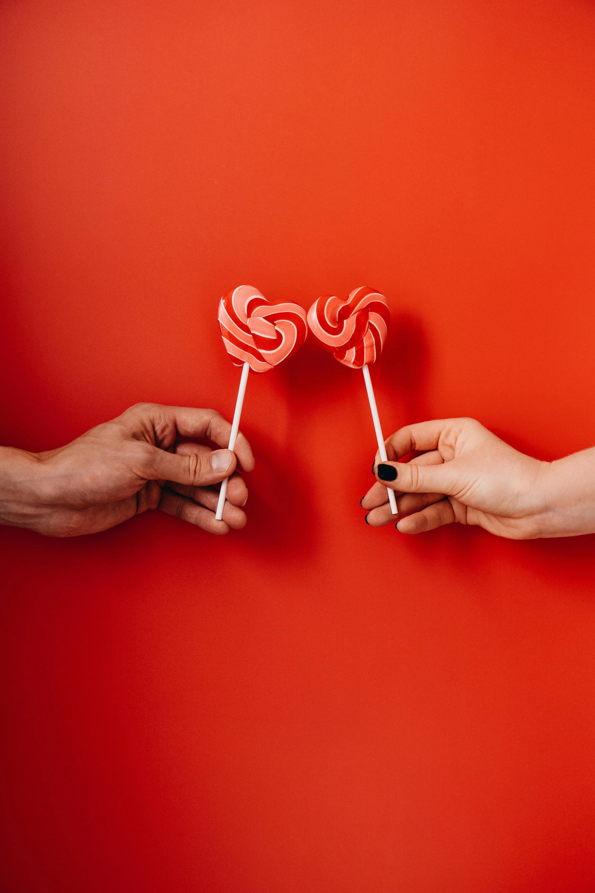 Two hands holding red and white swirled heart-shaped lollipops against a bright red background.