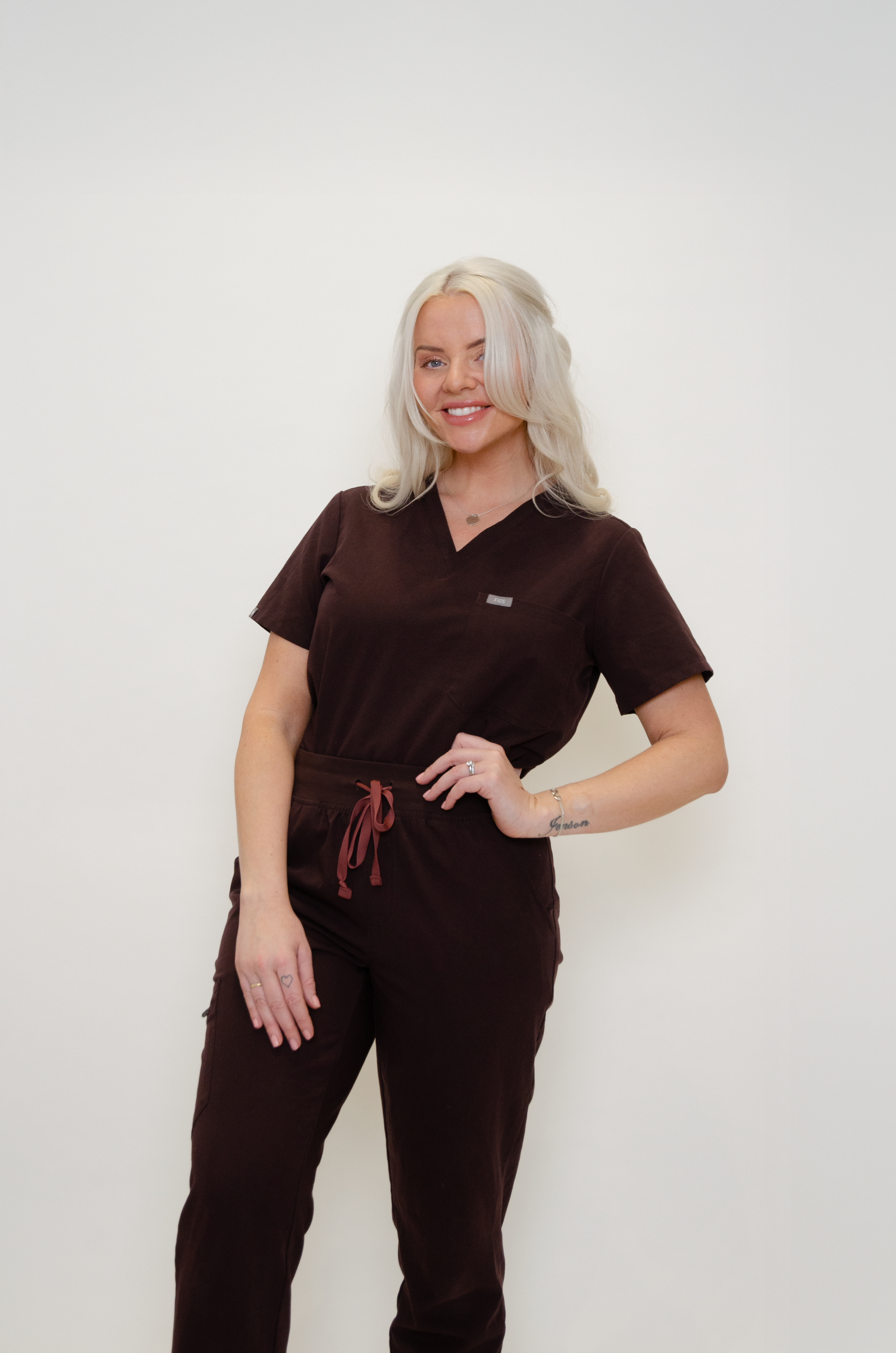 A woman with platinum blonde, shoulder-length hair, wearing a dark brown medical uniform, standing against a plain white background, smiling and posing with one hand on her hip.
