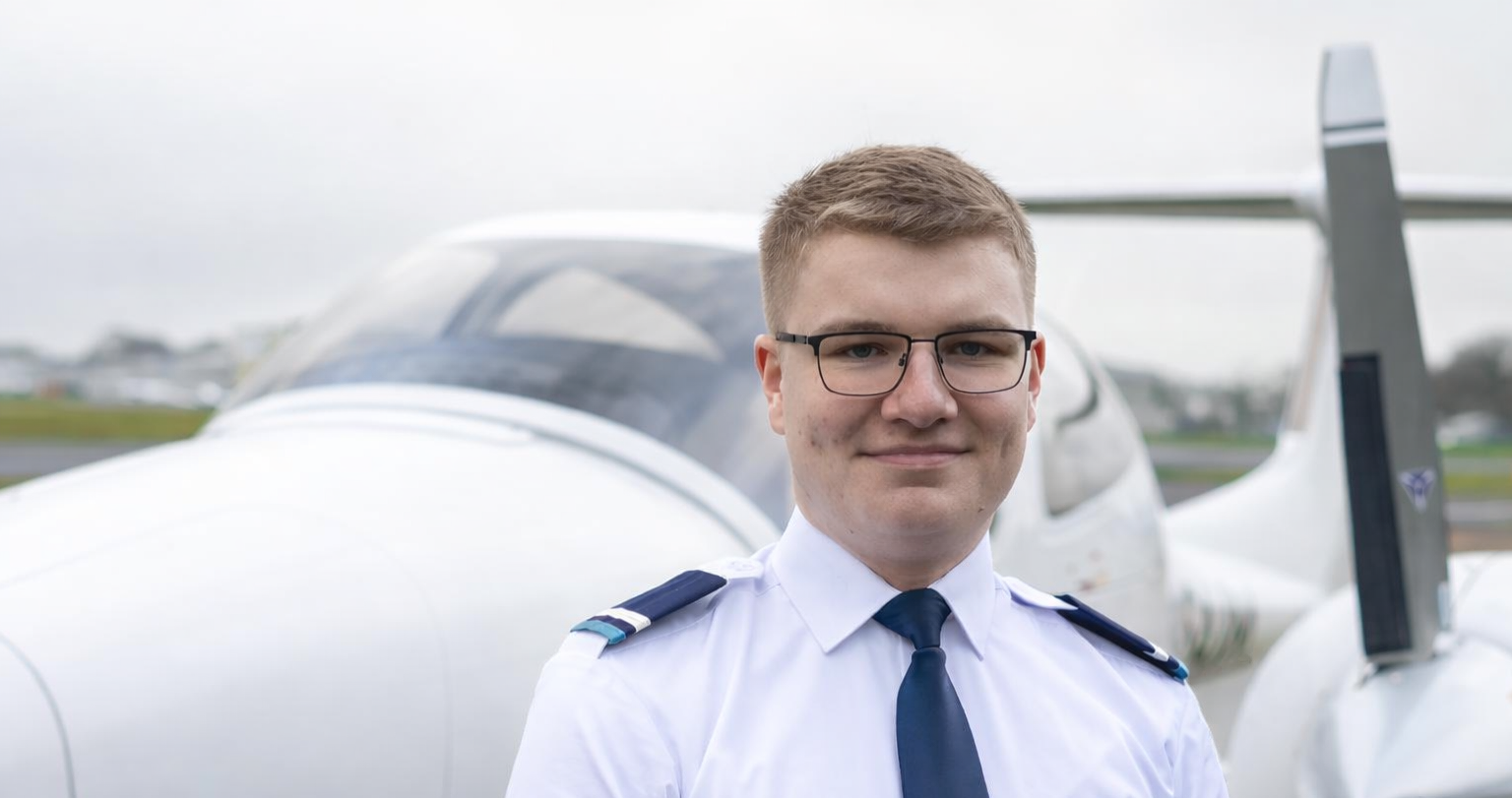 Young male pilot wearing glasses and a uniform with shoulder epaulets standing in front of a small white airplane.