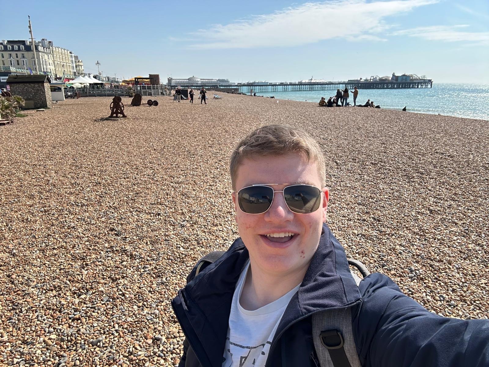 Smiling person wearing sunglasses and a jacket taking a selfie on a pebble beach with a pier and buildings in the background