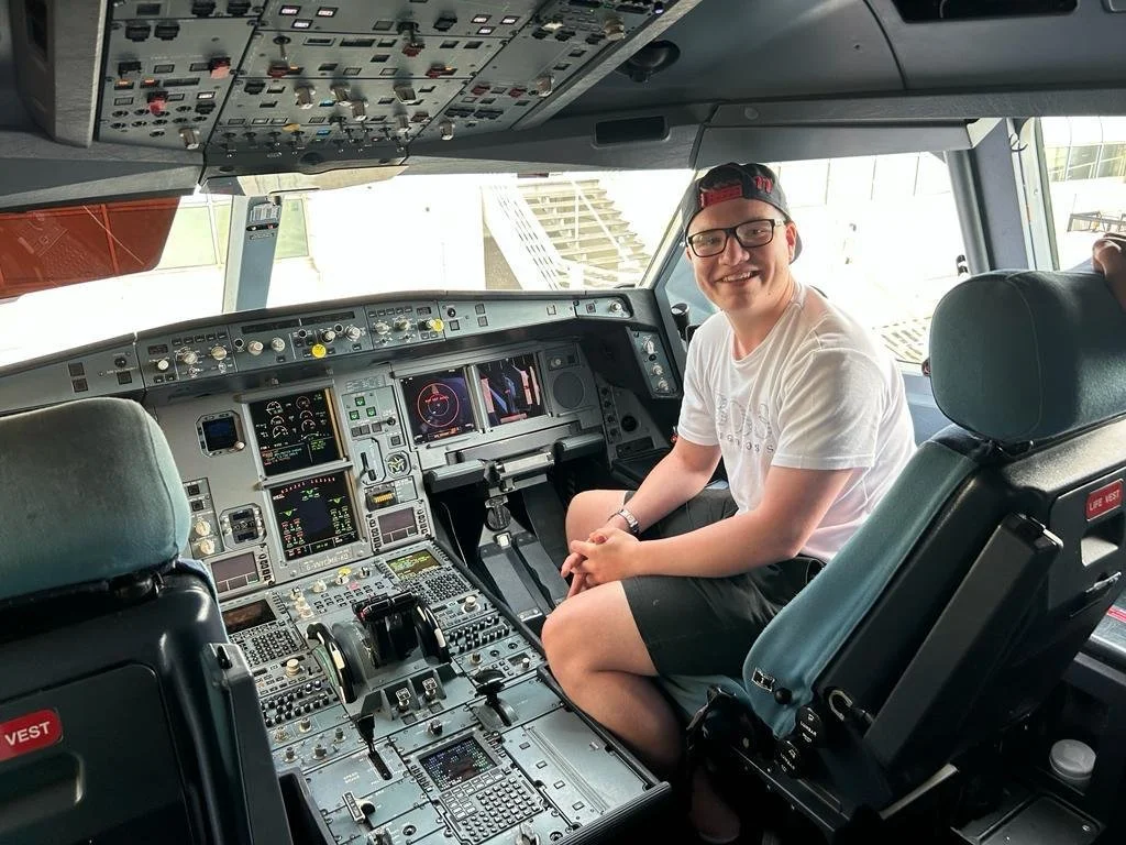 A young man sitting in the cockpit of an airplane, smiling at the camera, with a control panel and cockpit instruments visible.