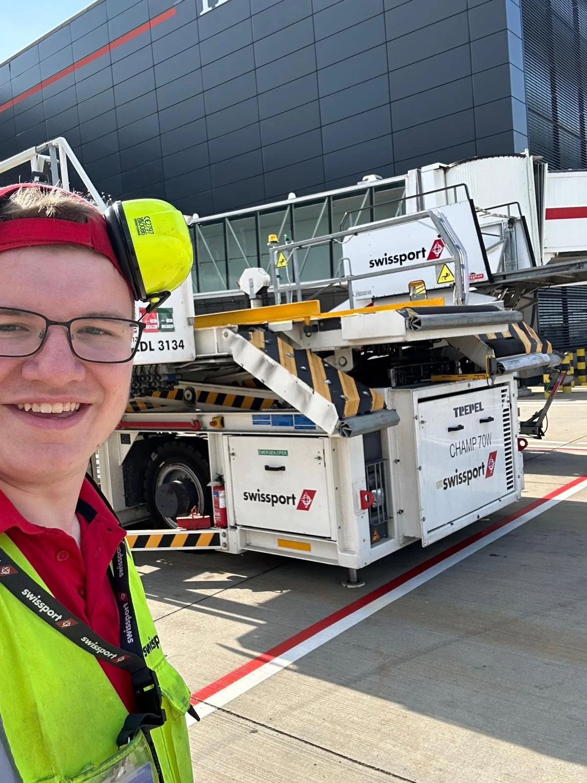 A smiling airport ground crew member wearing safety glasses, a red cap, and a neon yellow safety vest with a red shirt underneath. The crew member has yellow ear protection around the neck. Behind them is a mobile aircraft boarding staircase labeled 'TREPPEL CHAMP 70W' and 'swissport', positioned on an airport tarmac near a modern building.