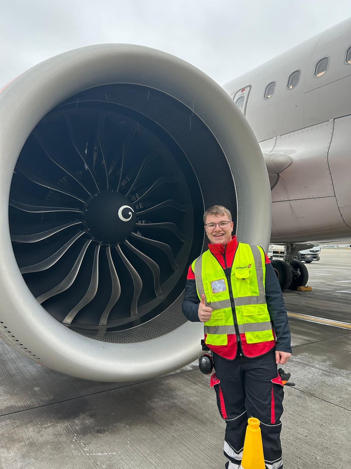 A smiling man wearing a high-visibility vest giving a thumbs-up next to a large airplane jet engine on the tarmac.