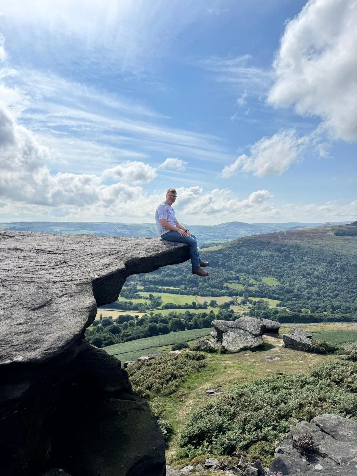A person sitting on the edge of a large rock formation overlooking a green valley with rolling hills and a partly cloudy sky.