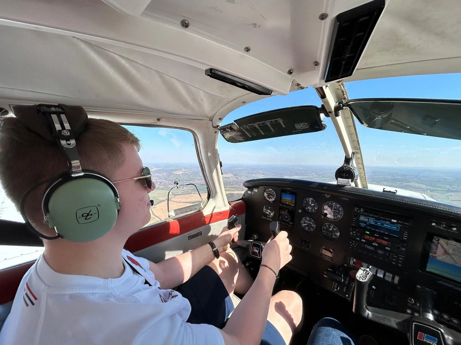 Young male pilot flying a small aircraft, wearing sunglasses, a white shirt, and large green noise-canceling headphones, with a view of the landscape below through the cockpit windows.