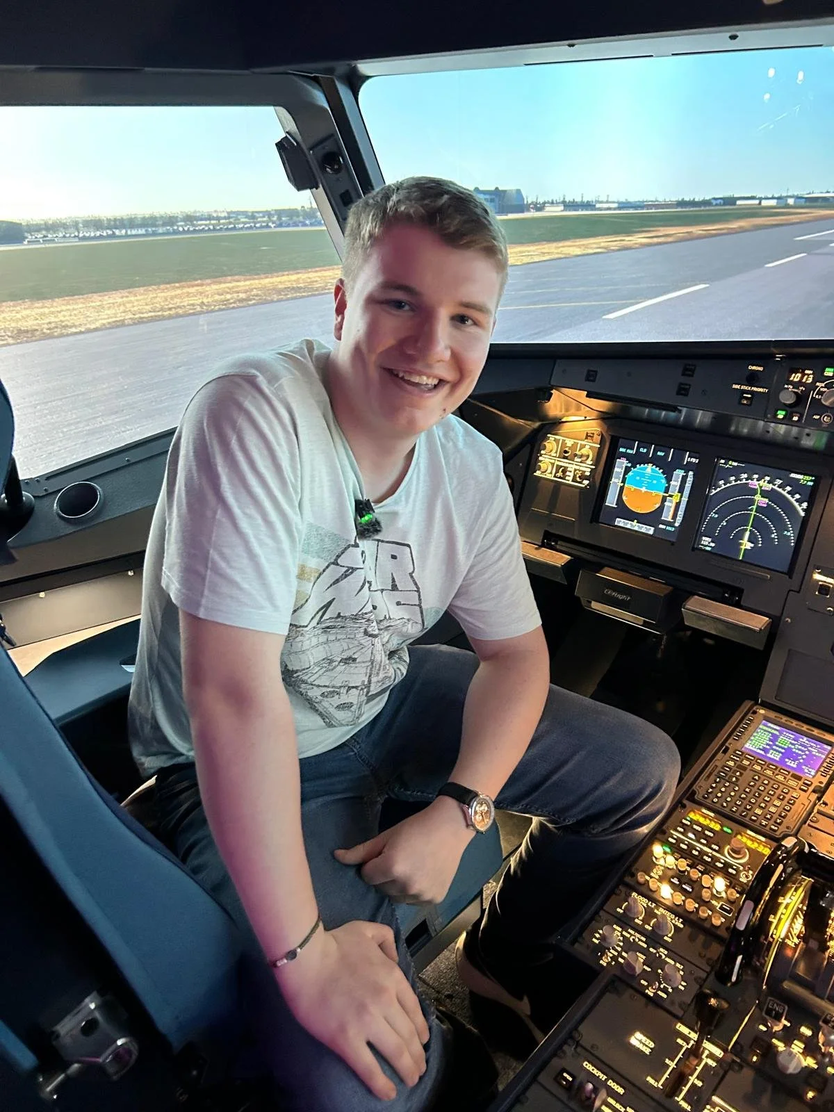 A young man sitting in the cockpit of an airplane, smiling at the camera with runway visible through the windshield behind him.